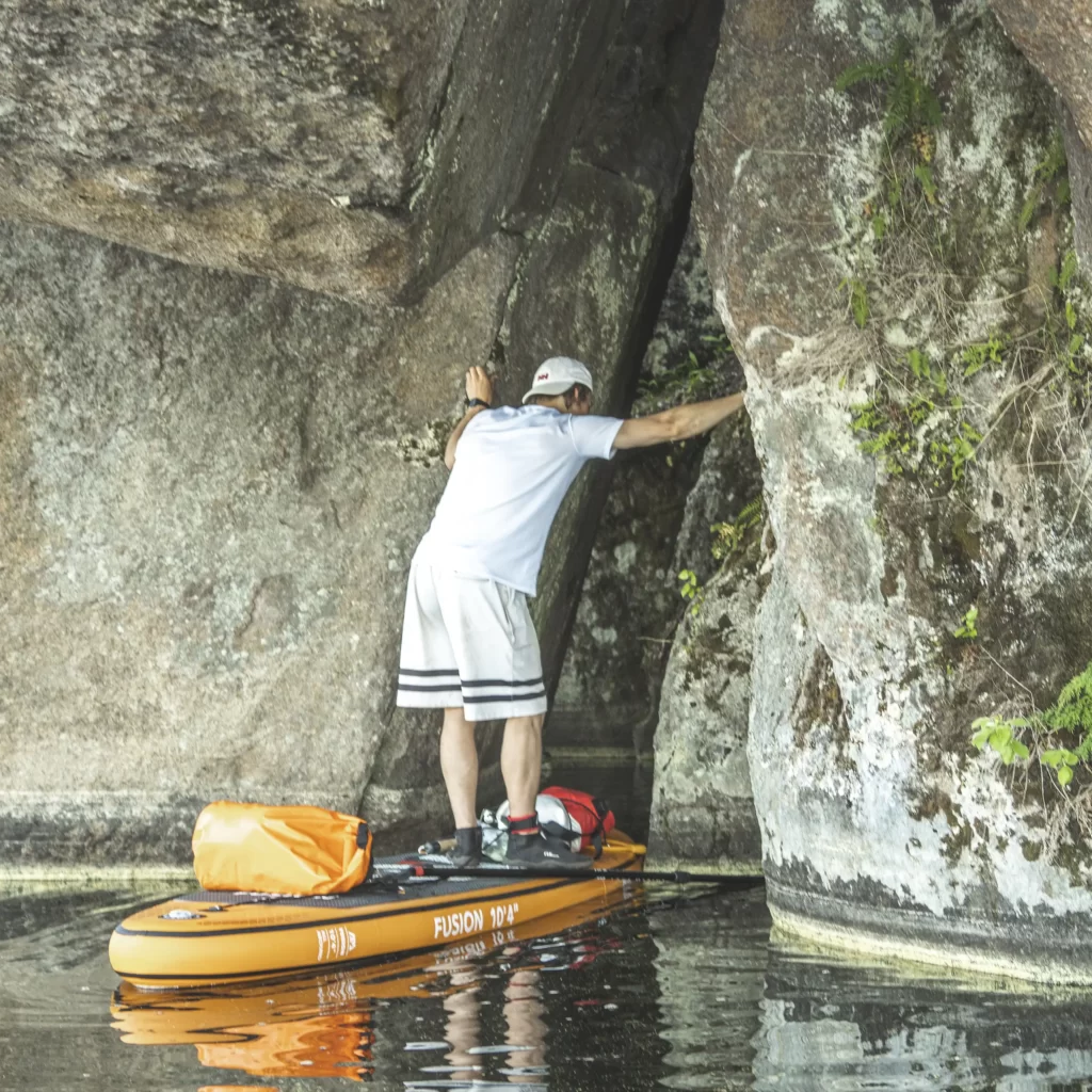 Mies nojailee kallioon sup-laudalla seisten vedessä kallionsuun luona