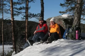 Mies ja nainen istuvat auringossa lumisessa maisemassa kahvilla